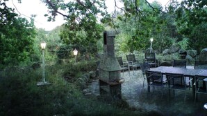 An outdoor dining area with stone tables and chairs, surrounded by lush greenery and a stone chimney structure. - Ferienpark Canow (Wustrow)