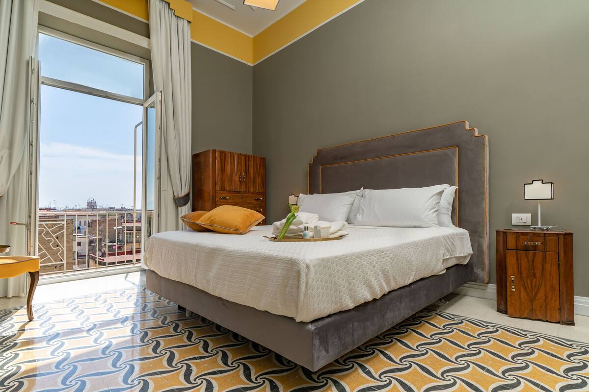 A bedroom with a large bed, a wooden wardrobe, a nightstand, and a view of the city through a window.