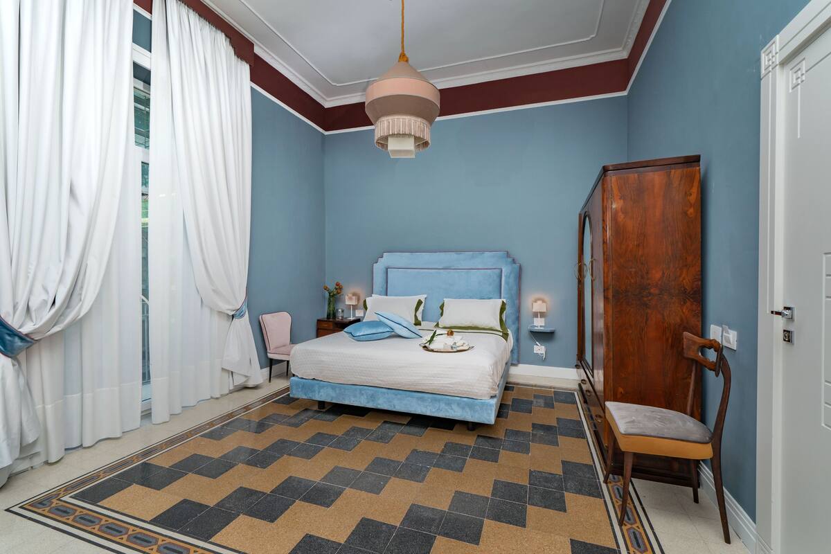 A bedroom with a bed, a wooden wardrobe, a chair, a nightstand, and a hanging light fixture.