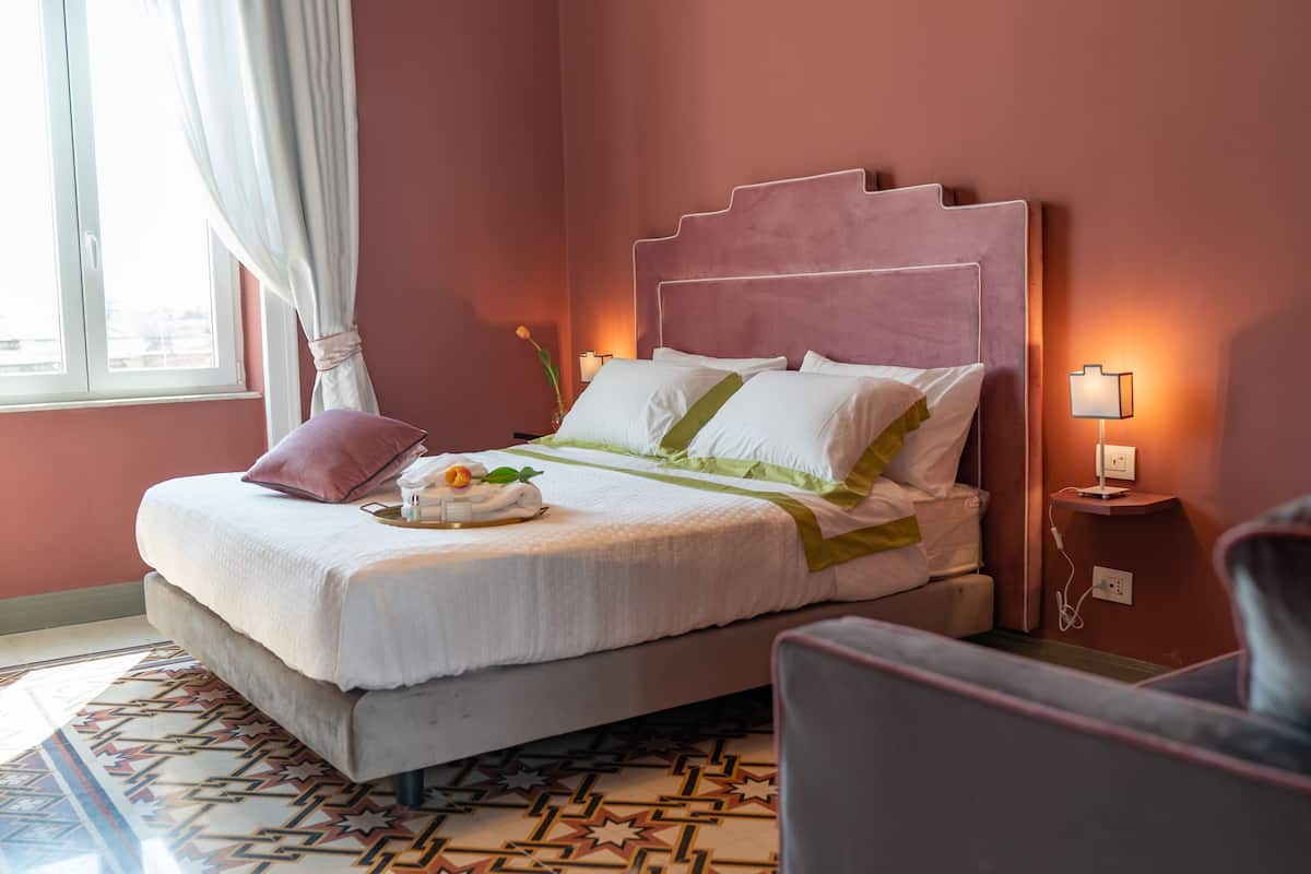 A neatly made bed with a headboard, a bedside table with a lamp, and a chair in a room with a patterned rug and a window with curtains.