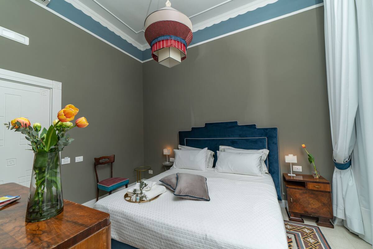 A neatly arranged bedroom with a bed, bedside tables, a nightstand, a vase with flowers, and a ceiling light fixture.