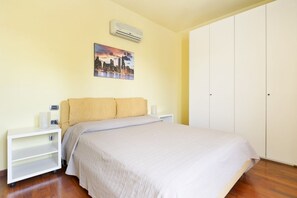 Une chambre à coucher comprenant un lit, une table de chevet, un climatiseur fixé au mur et un tableau représentant un paysage urbain.