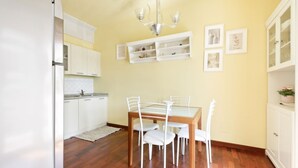 Un'area pranzo con tavolo in legno e sedie bianche, una cucina con mobili bianchi e un lampadario.