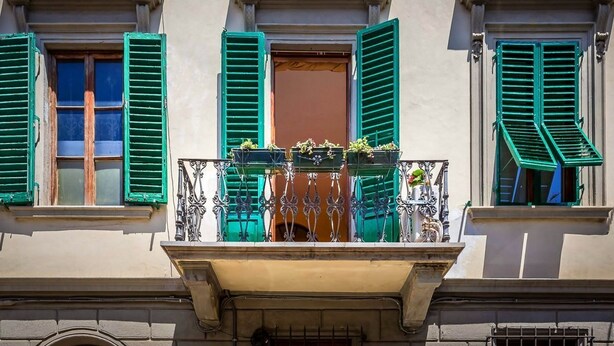 Ein Gebäude mit drei Fenstern, jedes mit grünen Fensterläden, einem Balkon mit schmiedeeisernen Geländern und Blumenkästen an den Fensterbänken.