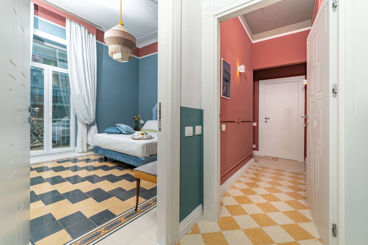 Une chambre à coucher avec un lit, une fenêtre avec des rideaux, un luminaire suspendu et un sol à carreaux.