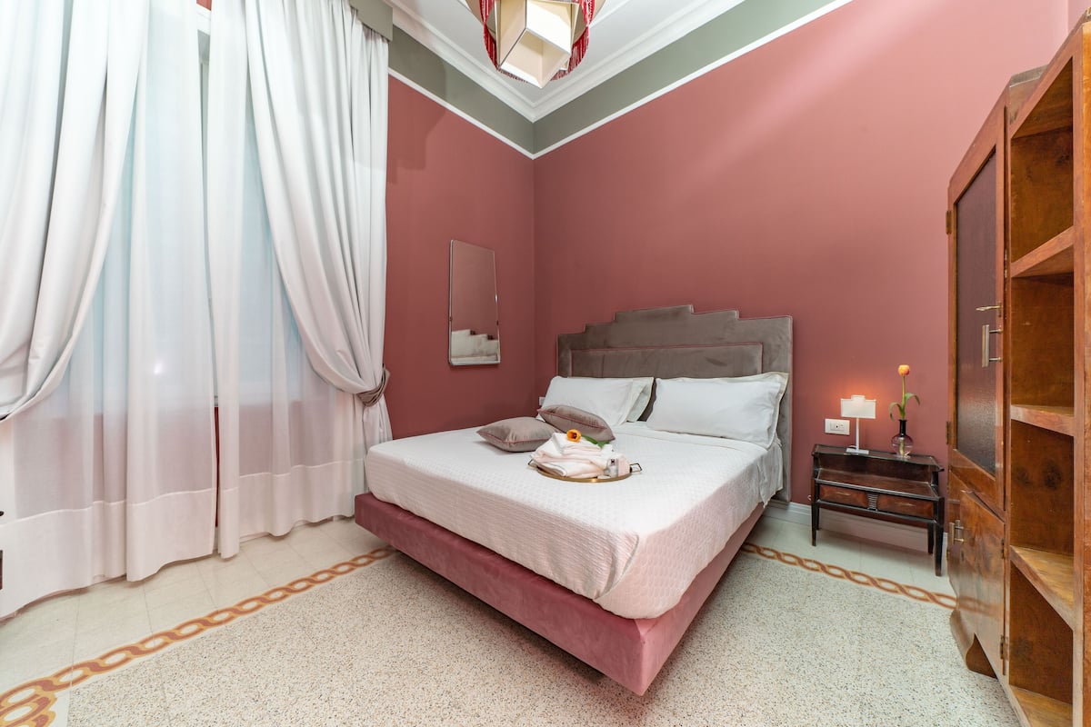 Un lit bien fait, avec une tête de lit rose, une table de chevet avec une lampe et une armoire en bois.