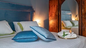 Una cama bien hecha con almohadas azules y blancas, una bandeja con toallas y una flor, y un armario de madera con espejo.