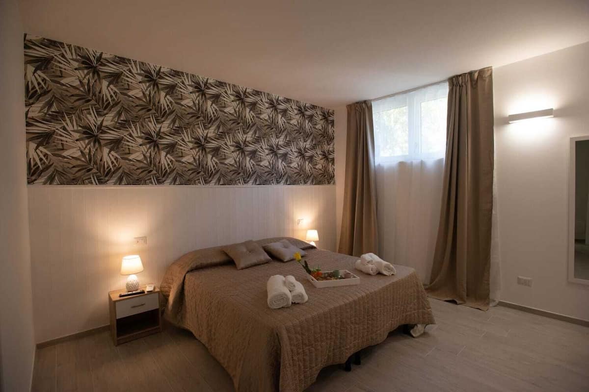 Un dormitorio con cama, mesitas de noche, una obra de arte en la pared y una ventana con cortinas.