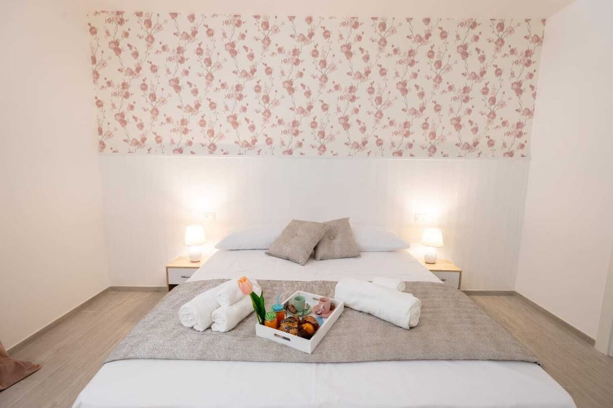 Un dormitorio ordenado con un cabecero estampado con motivos florales, una cama impecable con ropa de cama blanca, dos lámparas de noche y una bandeja con artículos de aseo.