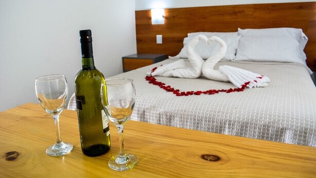 Une chambre d’hôtel avec une tête de lit en bois, un lit bien fait avec du linge de lit blanc et un élément décoratif rouge, ainsi qu’une table sur laquelle se trouvent une bouteille de vin et deux verres.