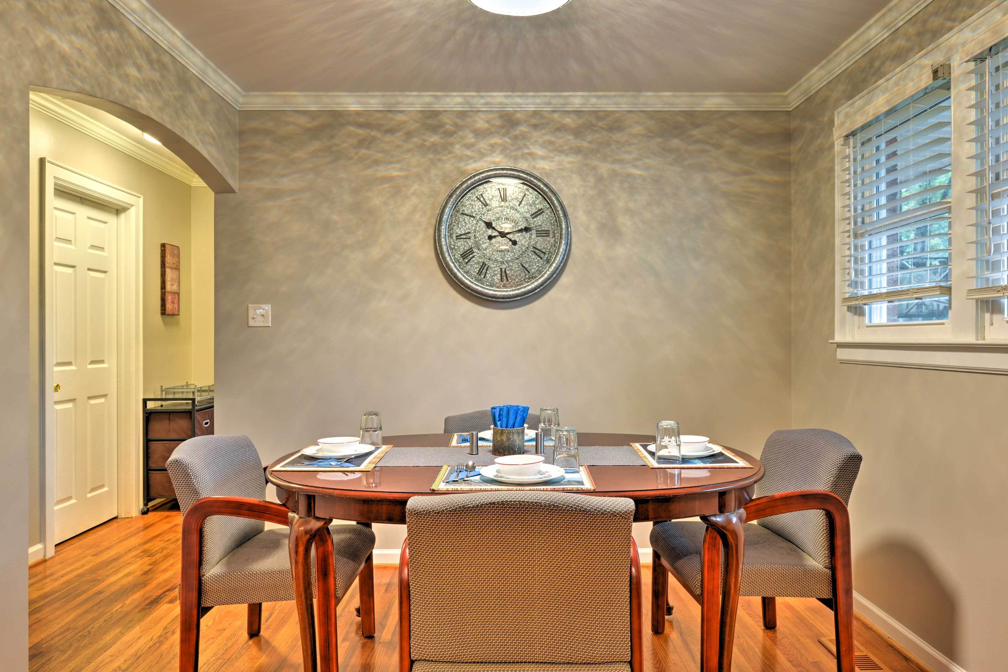 Une salle à manger avec une table ronde dressée pour quatre personnes, une horloge murale et une fenêtre avec des stores.