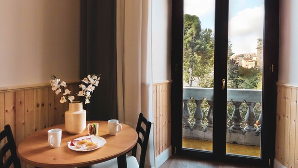 Uma área de refeições com mesa de madeira, cadeiras, um vaso com flores brancas e vista para uma varanda com grade e vegetação.