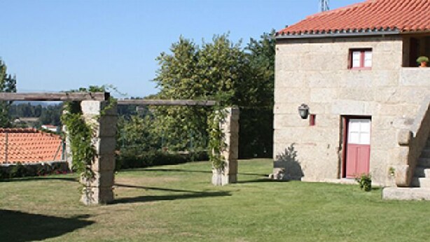 Ein Steinhaus mit roter Tür und kleinem Fenster, umgeben von einer gepflegten Rasenfläche und einer Pergola mit Kletterpflanzen.