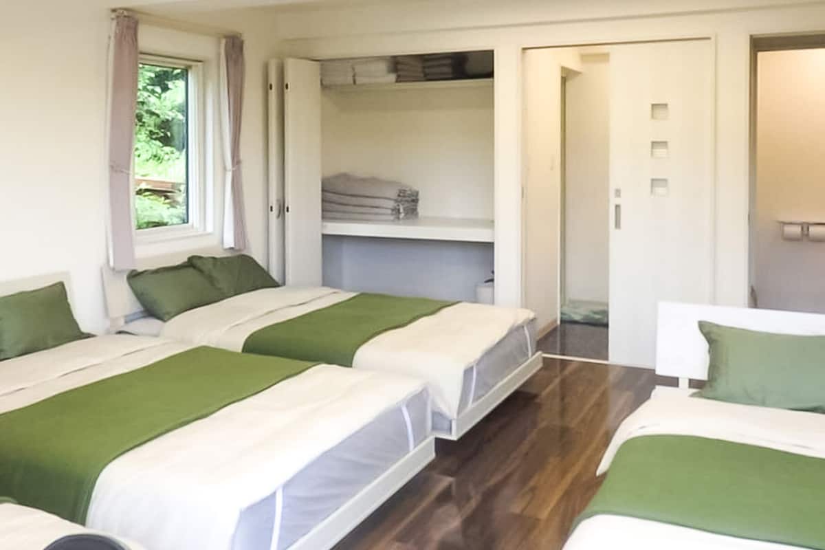 Una habitació amb tres llits, cadascun amb roba de llit verda i blanca. Hi ha una finestra amb cortines, una porta que dóna a un bany i una estant amb tovalloles doblegades.
