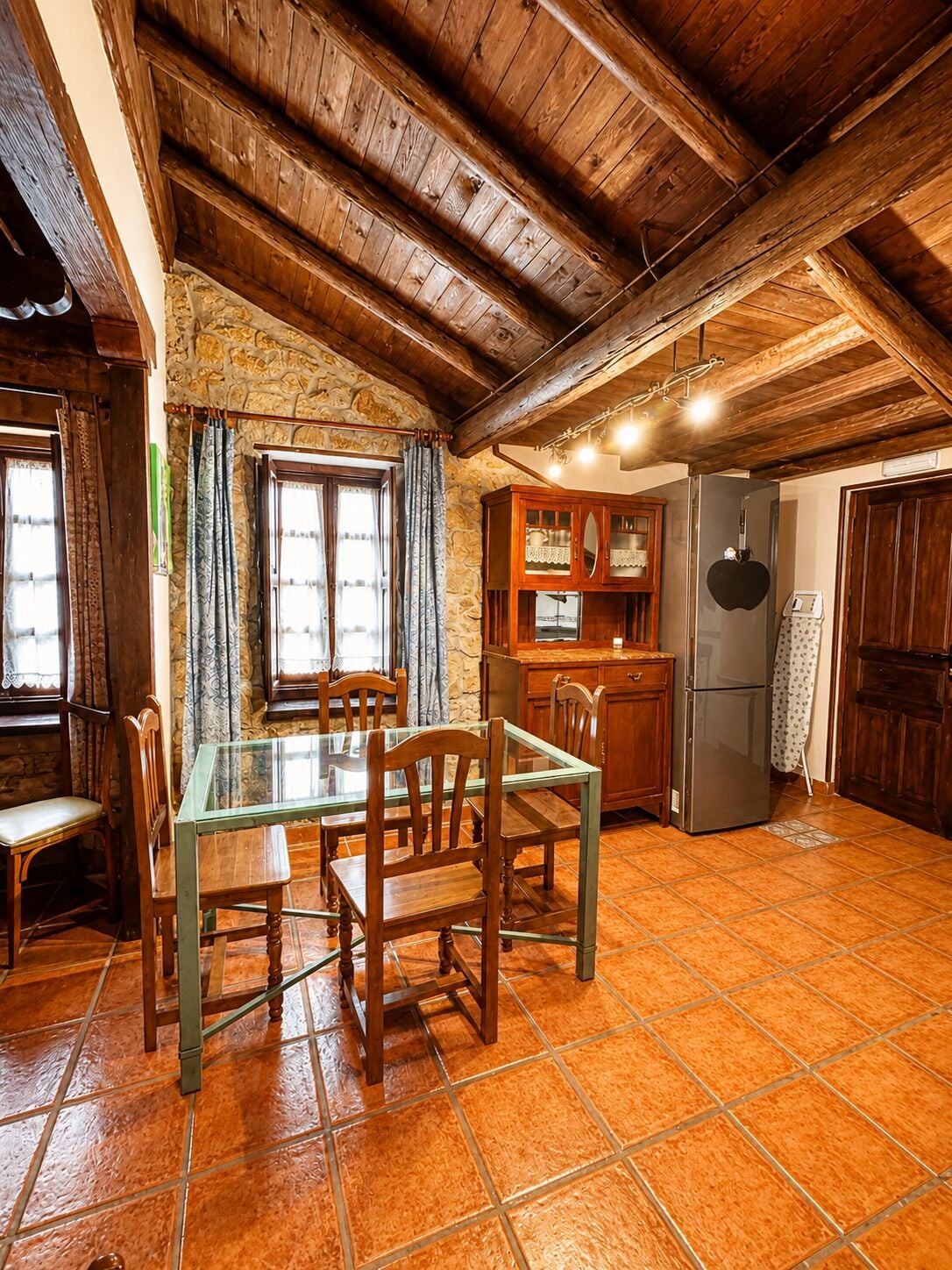 Una cocina rústica con pared de piedra, vigas de madera en el techo, una mesa de comedor con sillas, un refrigerador y una chimenea de piedra.