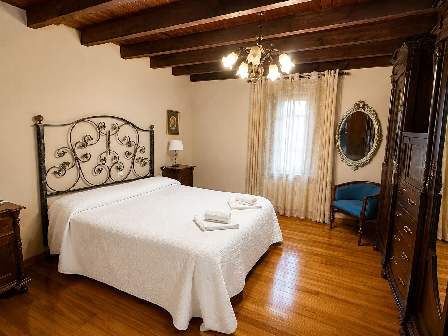 Une chambre à coucher avec un lit, un plancher en bois, un lustre, une chaise bleue, un miroir et une commode.