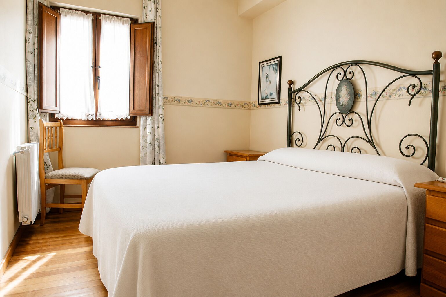 A bedroom with a bed, a chair, a wooden nightstand, a radiator, and a window with curtains.