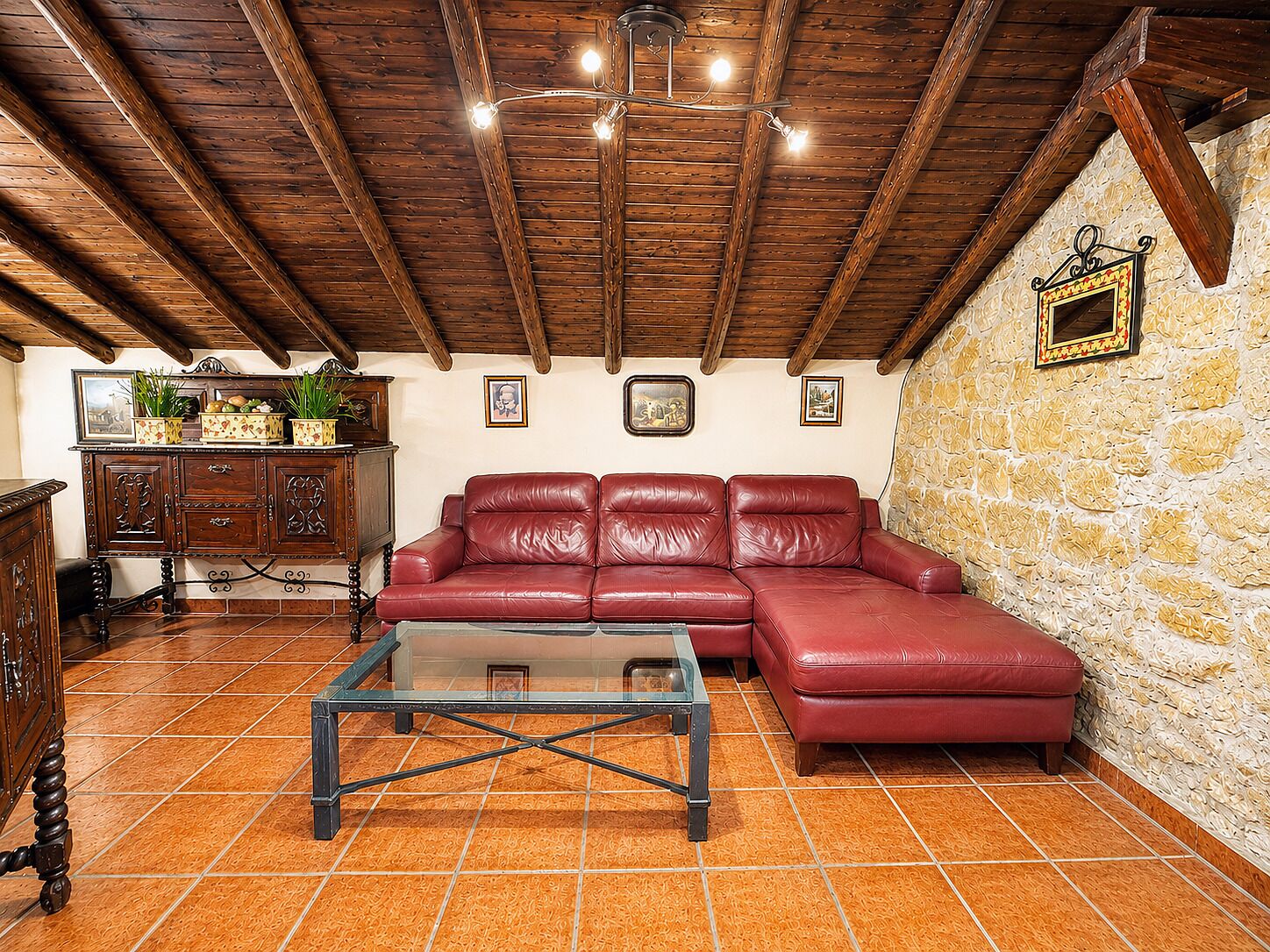 Una sala de estar con un sofá de cuero rojo, una mesa de centro de vidrio, vigas de madera en el techo y una pared de piedra.
