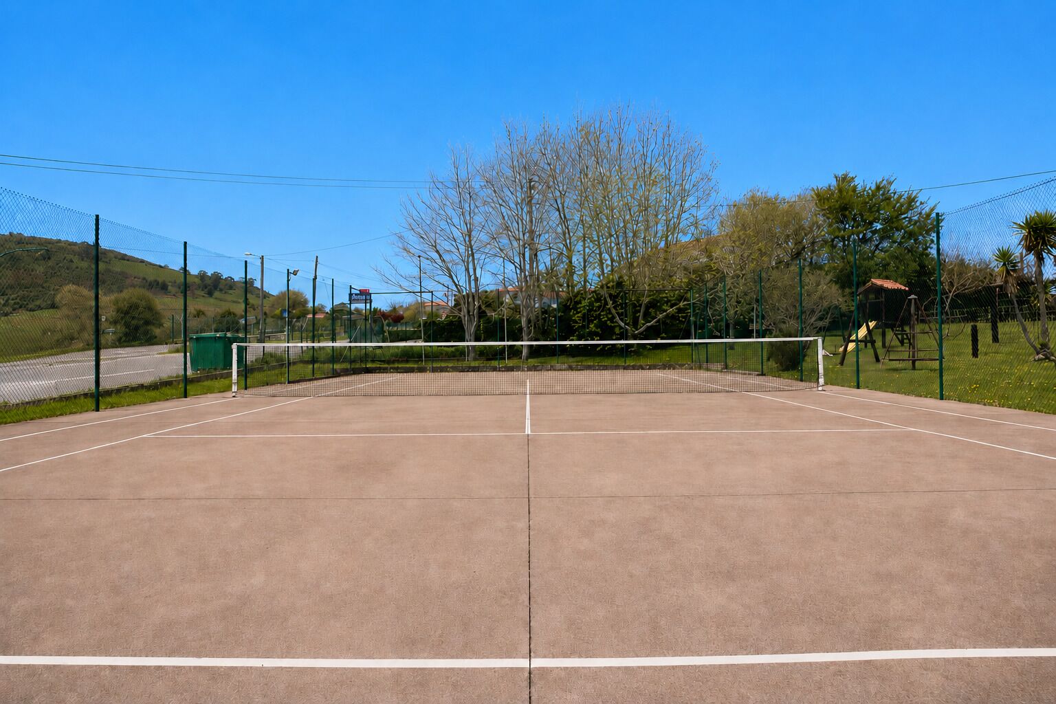 Een buiten tennisbaan met een net, omringd door een hek en bomen op de achtergrond.