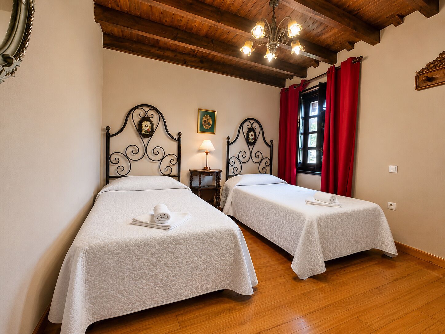 A room with two beds, wooden flooring, a chandelier, and a window with red curtains.