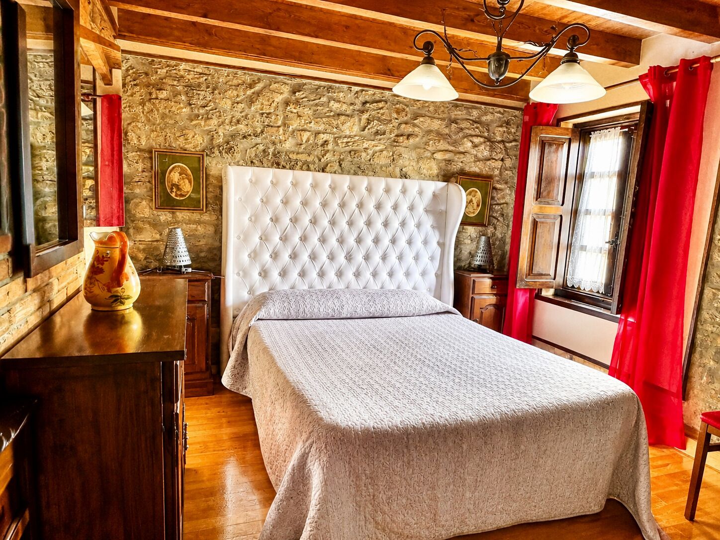 A bedroom with a tufted headboard, a wooden dresser, a red curtain, and a stone wall.