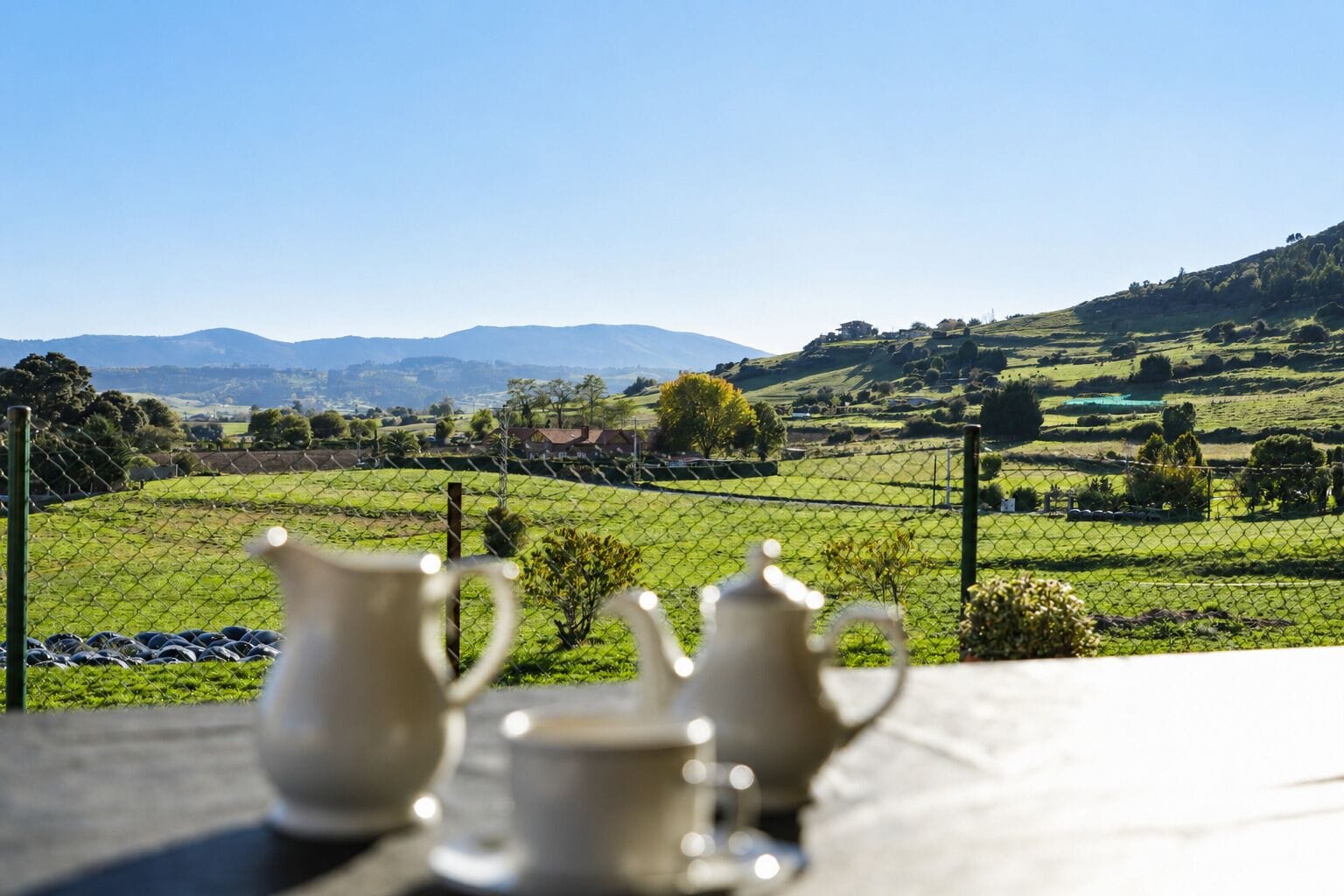 Une table sur laquelle sont disposés trois théières et trois tasses en céramique blanche, offrant une vue sur un paysage verdoyant avec des champs et des montagnes lointaines.