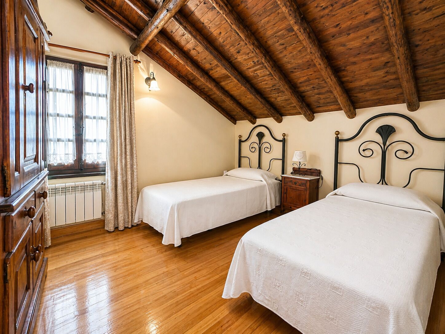 Una habitación con dos camas, piso de madera, radiador, ventana con cortinas y techo de madera.