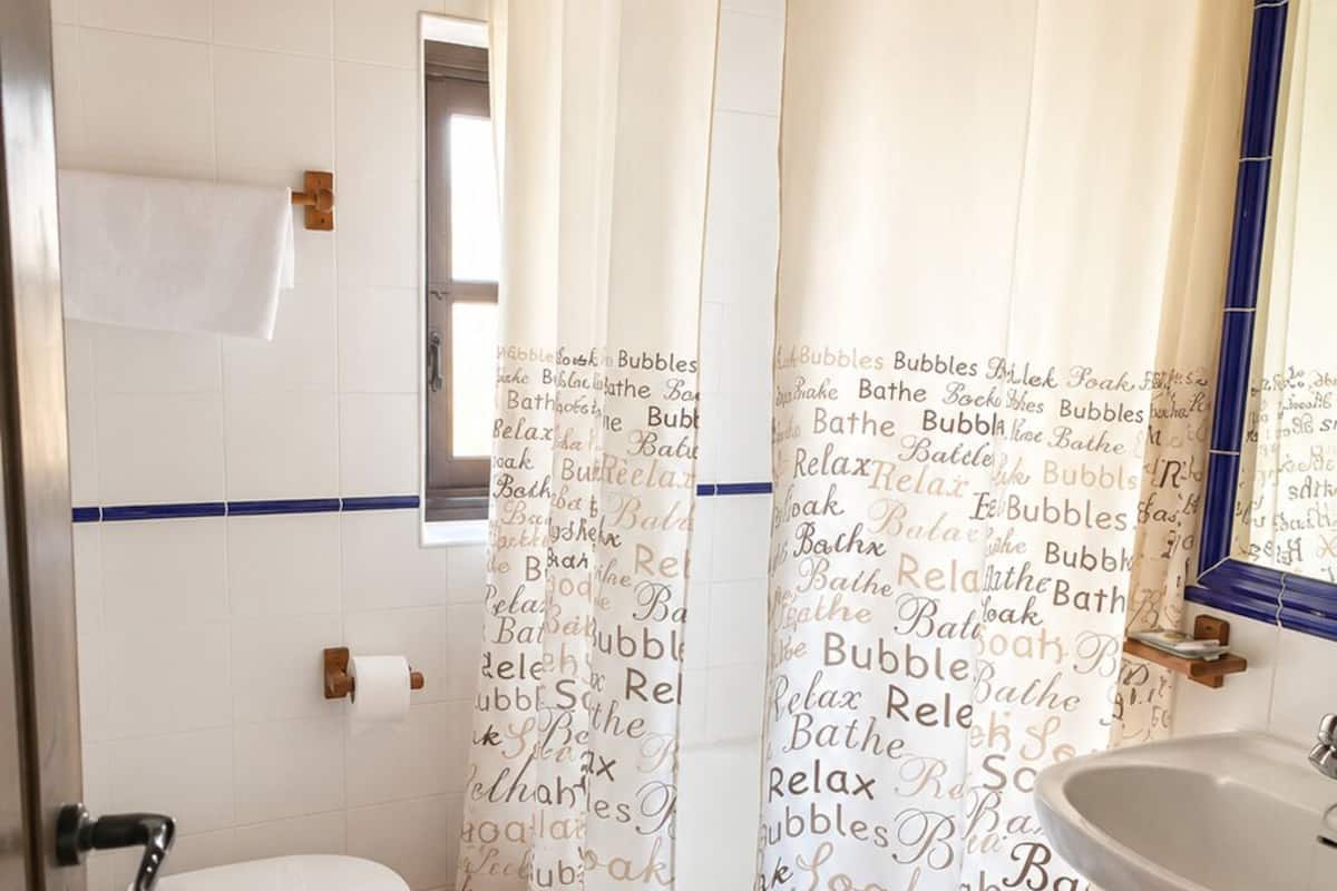 Casa de banho com sanita, bidé e lavatório, cortina de chuveiro com texto e vigas de madeira no teto.
