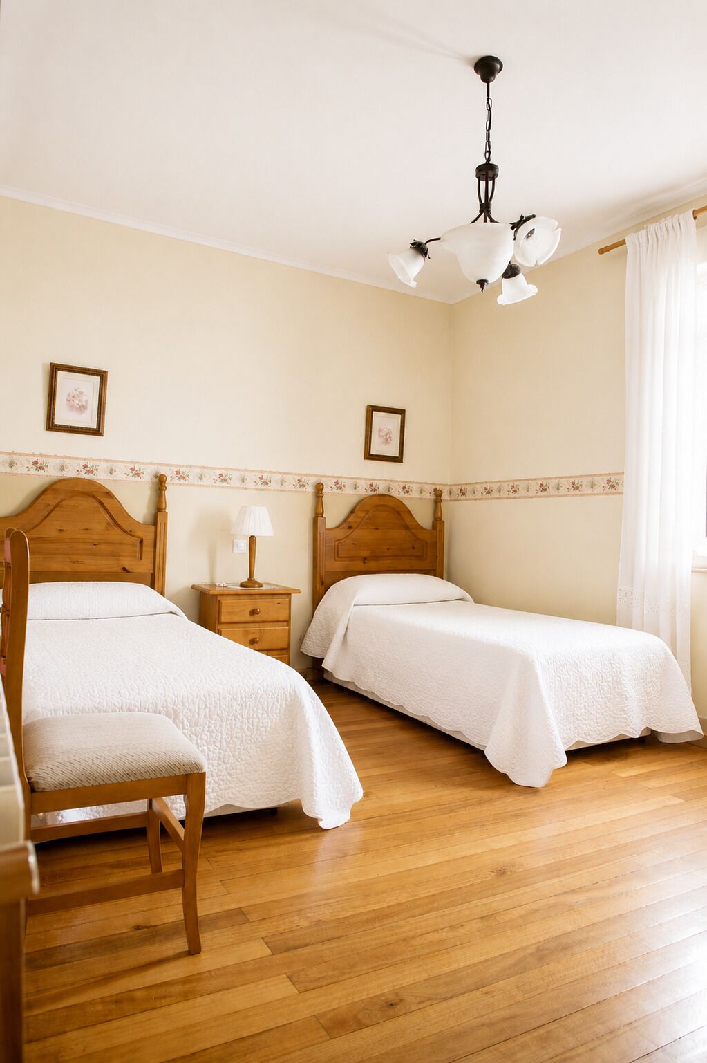 Una habitación con dos camas individuales, una silla de madera, una mesita de noche con lámpara y una lámpara de techo.