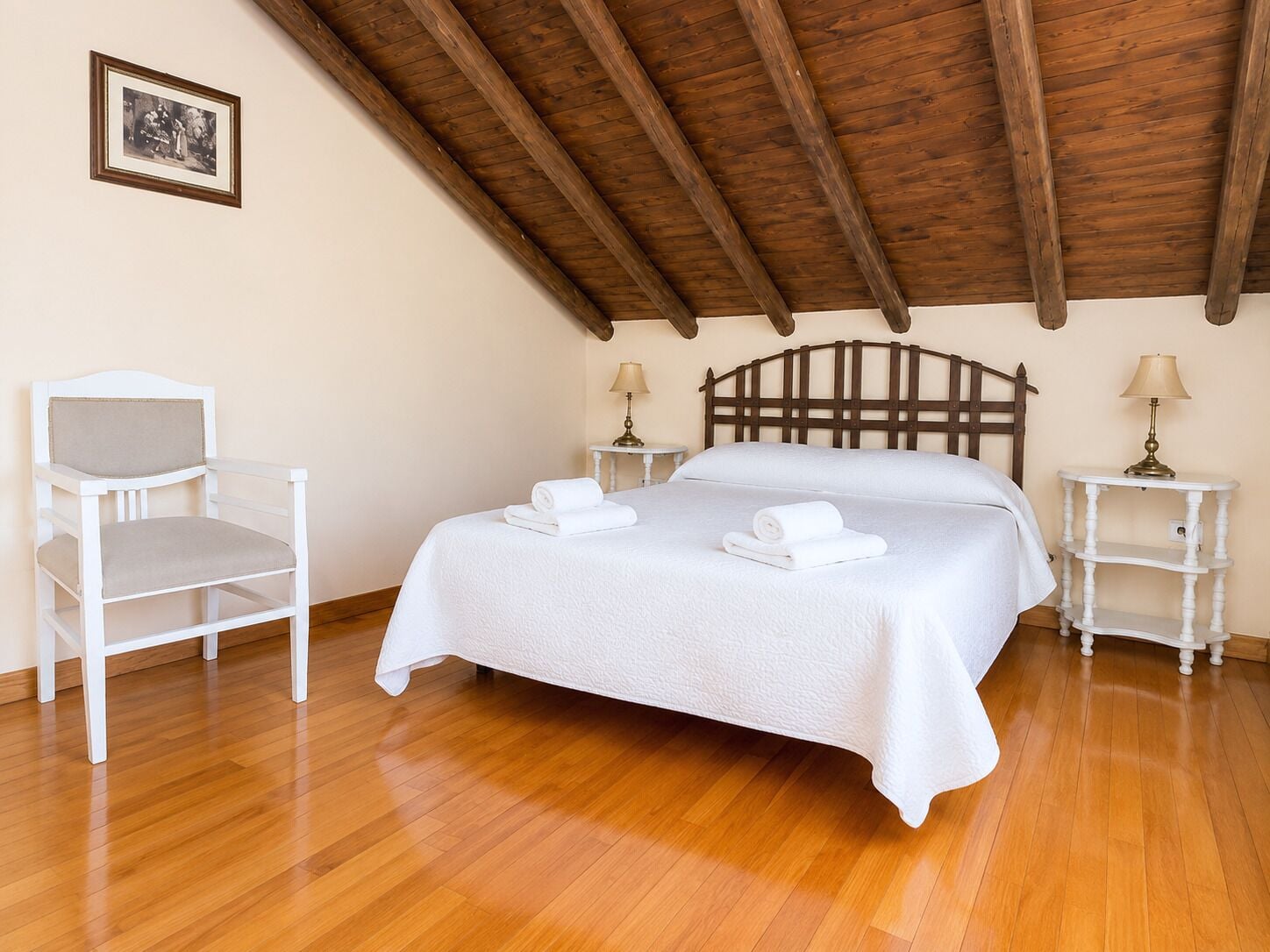 A bedroom with a wooden ceiling, a bed with white linens, a white chair, and bedside tables with lamps.
