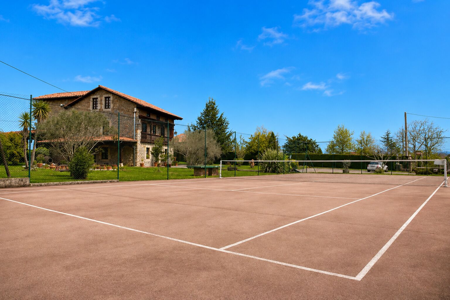 A tennis court with a net, surrounded by a fence, and a building with a stone facade in the background.