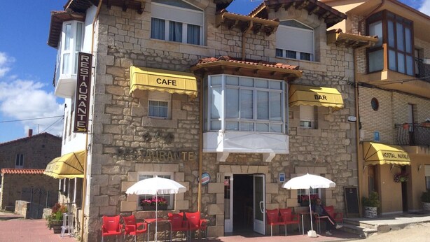 A stone building with a cafe and bar on the ground floor, outdoor seating with red chairs and white umbrellas, and a sign indicating a restaurant. - Hostal Mónica (Valle De Valdebezana)