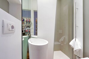 Un baño moderno con un lavamanos blanco, una cabina de ducha con mampara de vidrio y una estantería visible al fondo.