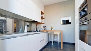 A modern kitchen with white cabinetry, a wooden countertop, and a built-in oven. There are two white bar stools and a small table with a bottle and glasses. - San Lorenzo (Rome)