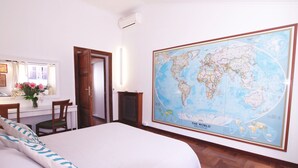 Una habitación de hotel con un gran mapa mundial en la pared, una cama con ropa blanca, un escritorio con una silla y una ventana con cortinas.