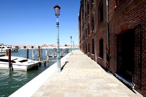 A waterfront walkway with a brick building on one side and a dock with boats on the other. - Giudecca Apartment View (Venice)