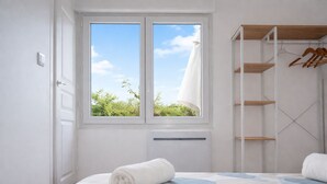 A bedroom with a bed, a window with a view of greenery, a radiator, and a wooden wardrobe with hanging space. - La Belle Aultoise, Ault center, sea view, Baie de Somme (Ault)