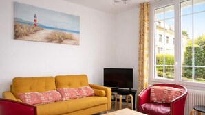 A living room with a yellow sofa, red armchair, and a TV. There's a painting of a lighthouse on the wall and a window with a view of buildings and greenery. - La Belle Aultoise, Ault center, sea view, Baie de Somme (Ault)