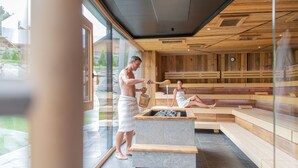 Una sauna amb parets de fusta i un banc de pedra, una persona abocant aigua i una altra relaxant-se.