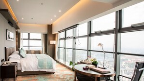 A modern hotel room with a large bed, a desk with a lamp, and a view of the city through large windows. - Ramada Plaza by Wyndham Nanxun (Huzhou)