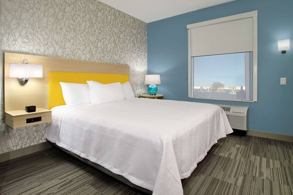 A hotel room with a bed, two bedside tables, a nightstand, a lamp, a globe, a window with blinds, and a wall-mounted air conditioner.