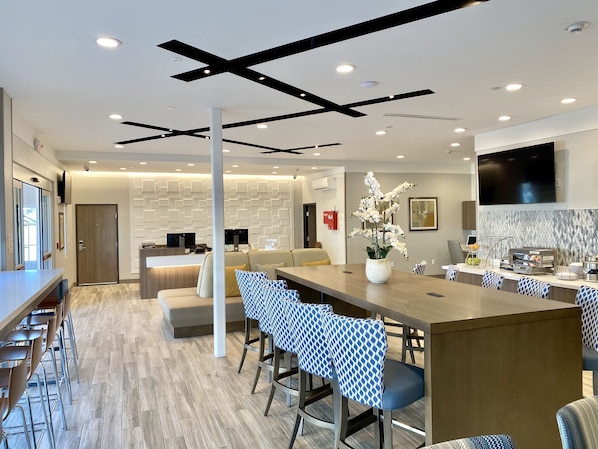 A modern hotel lobby with a long reception desk, seating area, and a kitchen in the background.