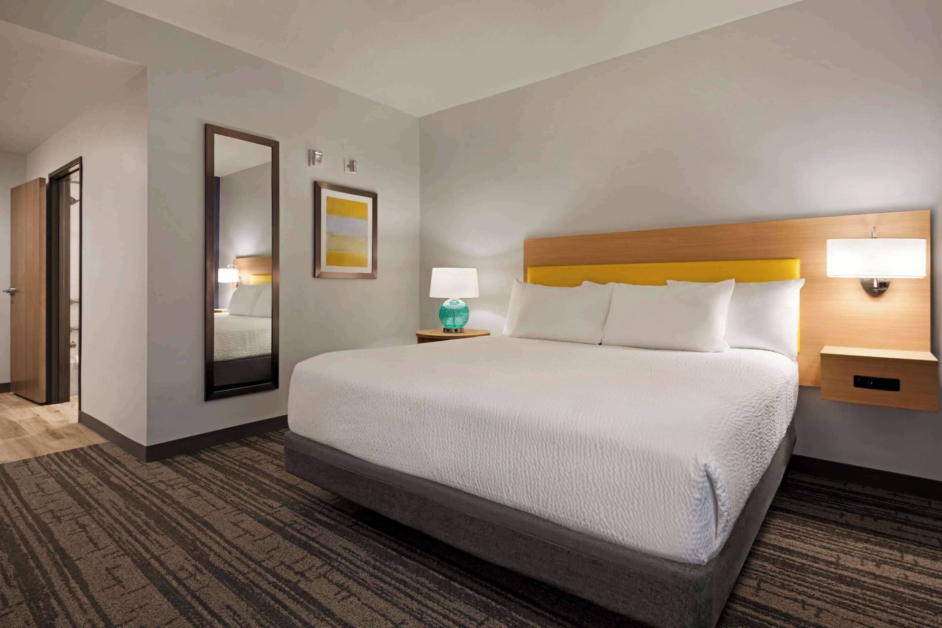 A hotel room with a large bed, a bedside table with a lamp, a mirror, and a picture on the wall.