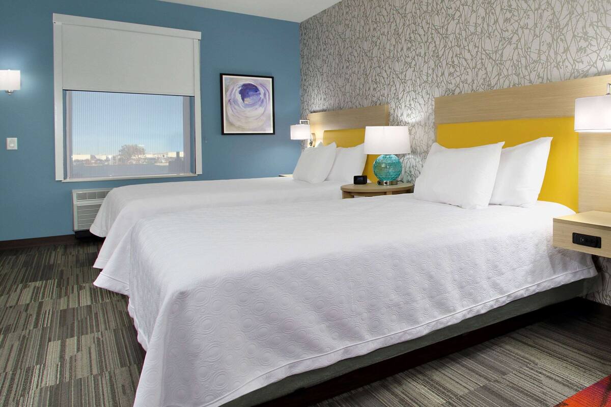 A hotel room with two beds, a large window, a wall-mounted artwork, and a patterned wallpaper.