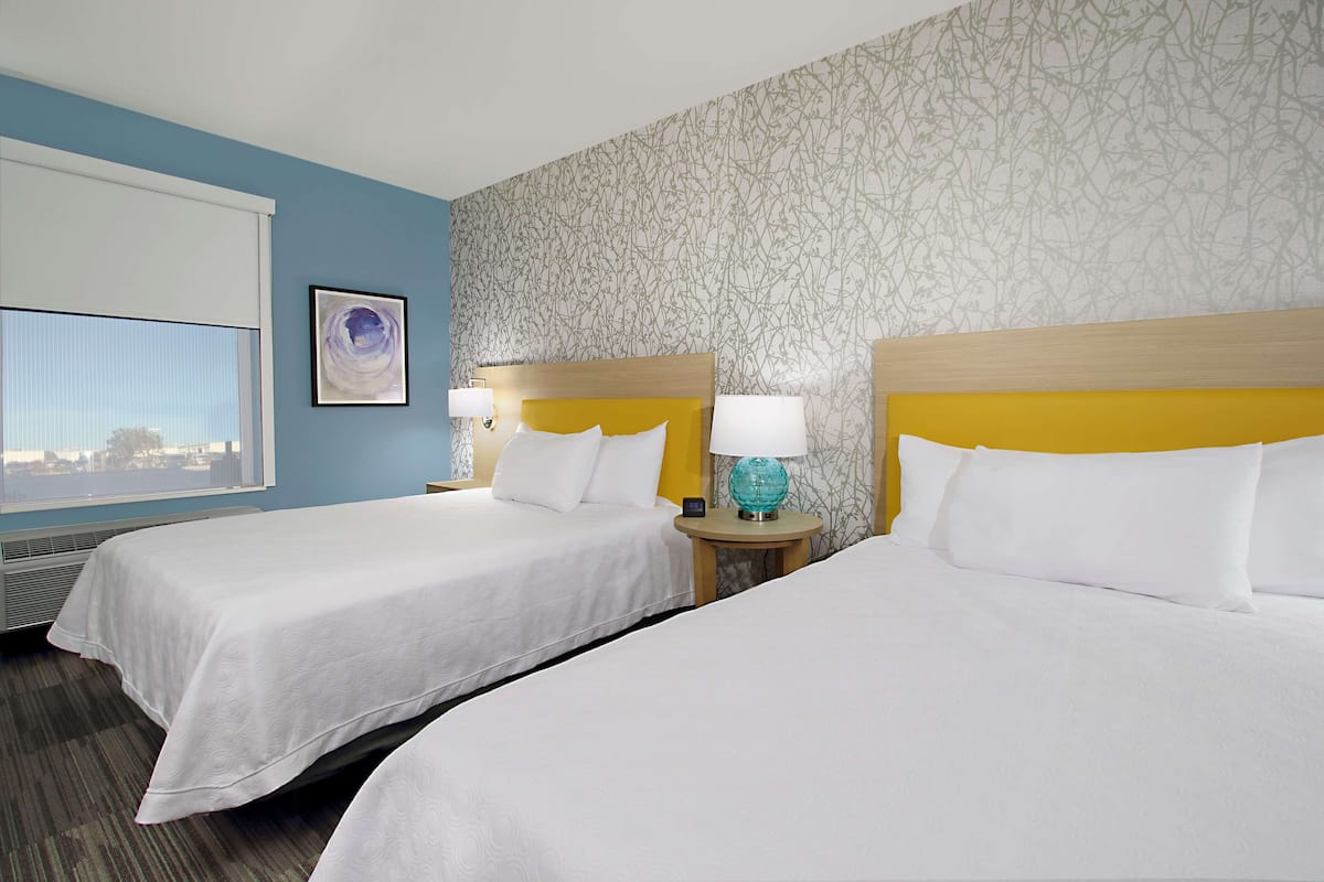 A hotel room with two beds, a window with a view, a patterned wall, and a framed picture on the wall.