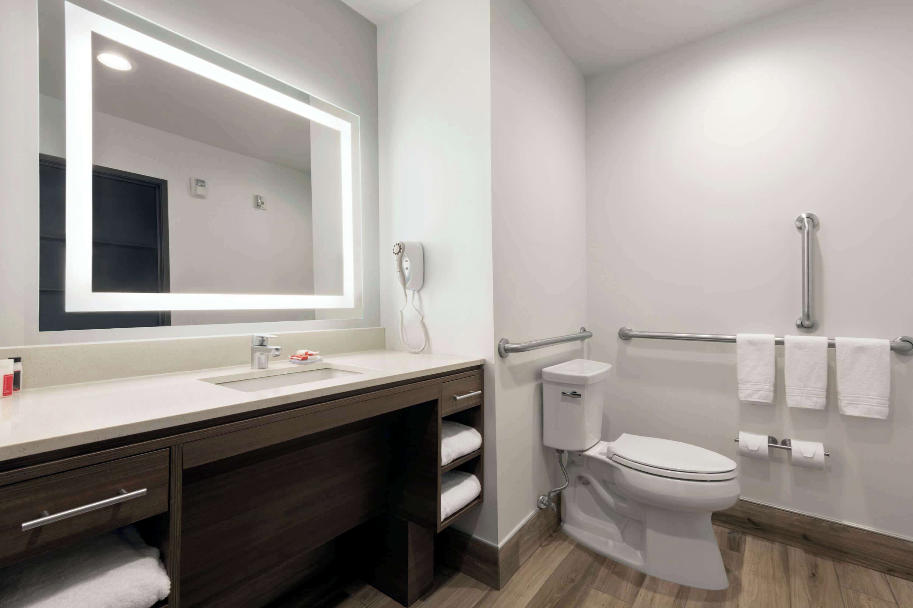 A modern bathroom with a large mirror, a sink, a toilet, and a handrail.