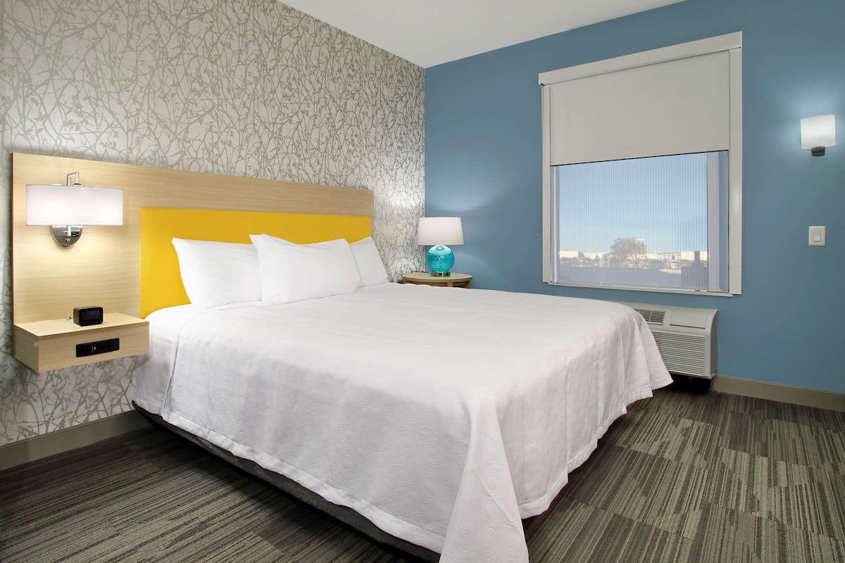 A hotel room with a bed, two bedside tables, a window with blinds, and a wall with a patterned wallpaper.