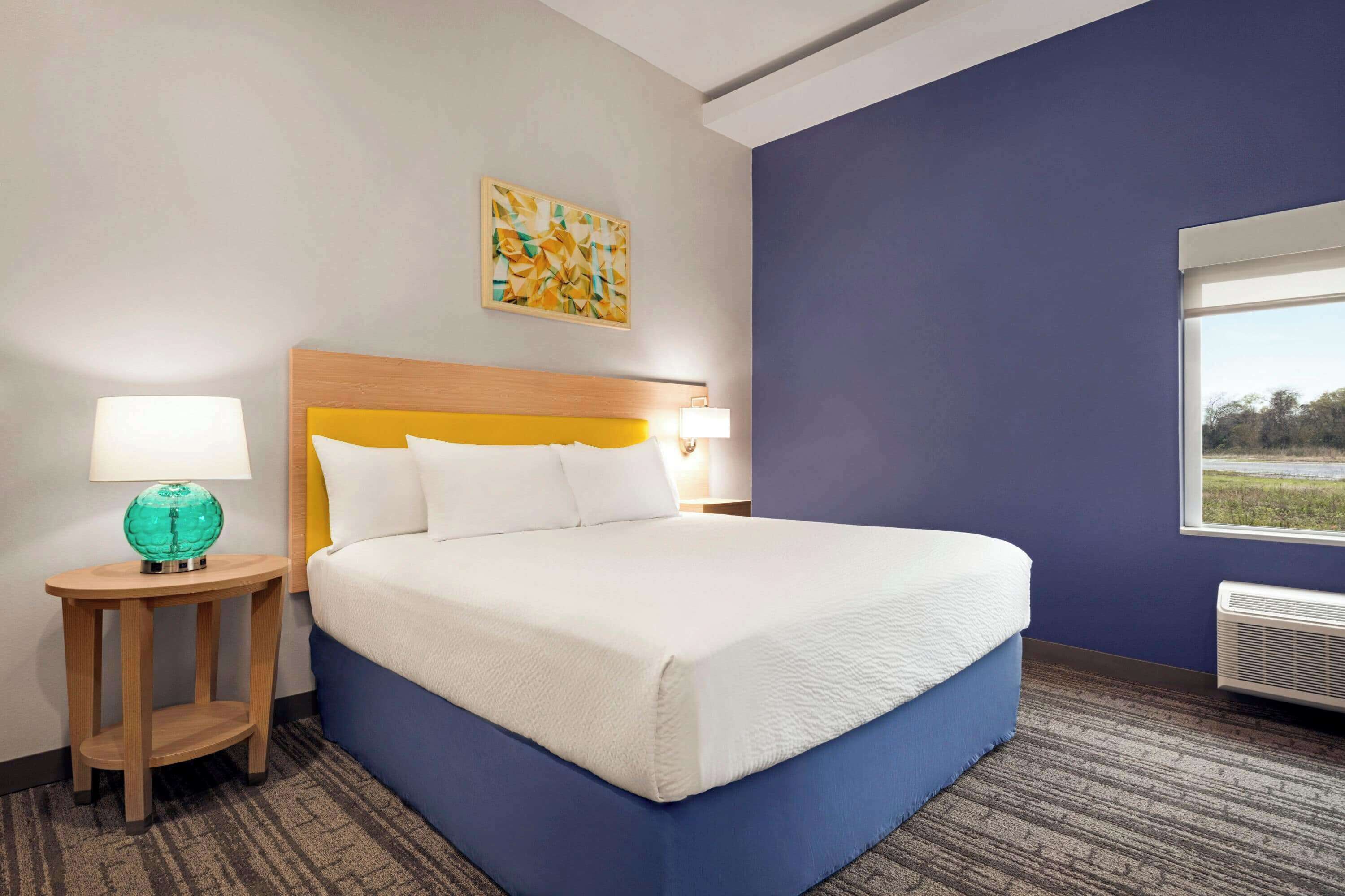 A hotel room with a large bed, a nightstand with a lamp, a painting on the wall, and a window with a view.