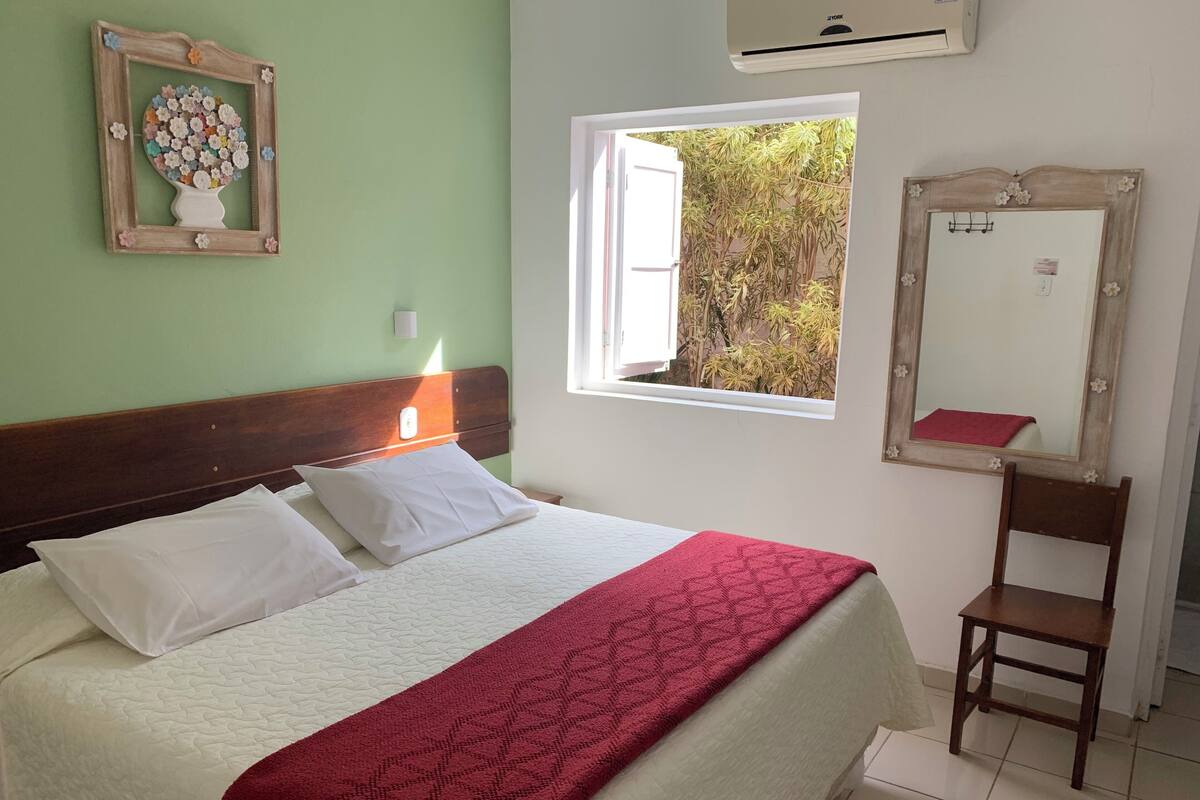 A bed with white bedding and a red blanket, a wooden headboard, a small bedside table, a wall-mounted mirror, and a window with a view of greenery.