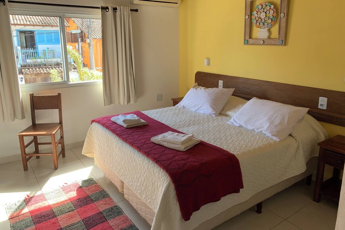 A neatly made bed with white linens, a red blanket, and a wooden headboard. There is a small wooden chair and a colorful checkered rug on the floor.